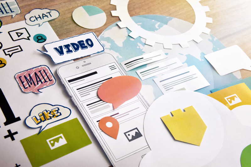 Paper cutouts of video, email, and other marketing terms in bubbles, cluttered on table with other marketing materials and cutouts