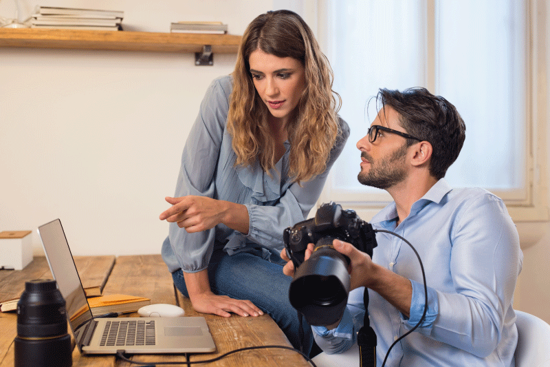 Man holding camera that is plugged into laptop next to woman pointing to laptop screen