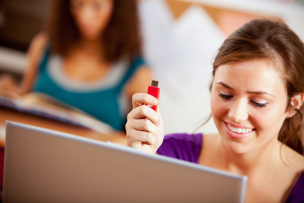 girl in purple shirt holding red flash drive while looking at computer screen